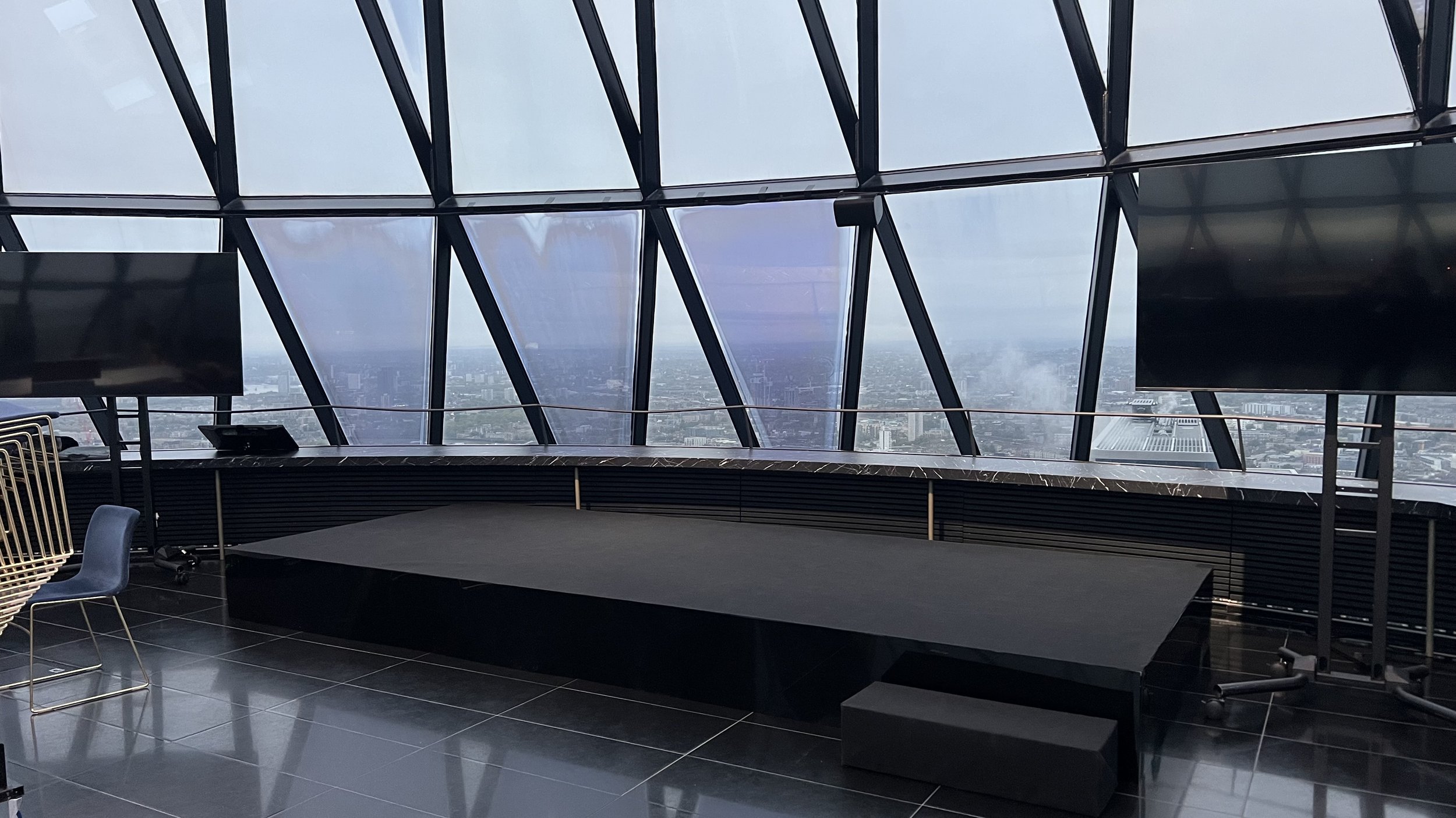 Interior view of a modern building with large glass windows showing a cityscape, a black marble counter, and TV screens, with chairs nearby.