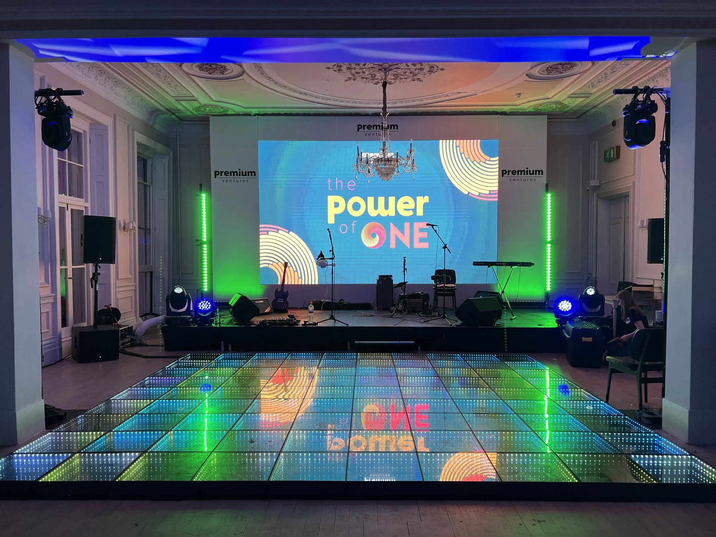 Event stage with a large LED screen displaying the text 'the power of ONE,' surrounded by colorful lights and musical instruments in a decorated room with a chandelier.