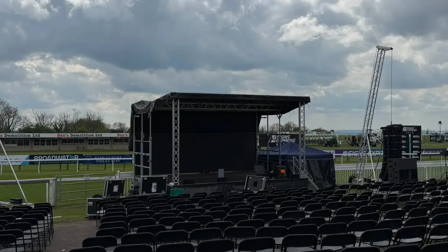 Empty outdoor stage setup with chairs facing a black stage, large speakers, and lighting equipment, under cloudy sky in a stadium or event field.