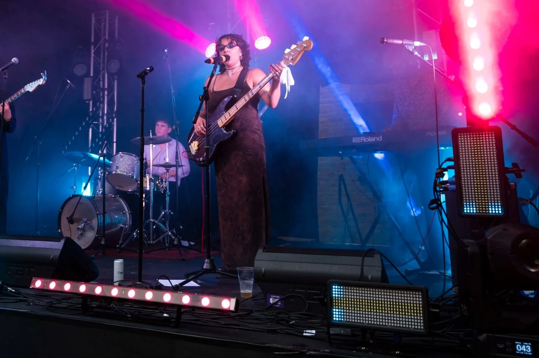 A female musician with curly hair, wearing sunglasses and a dark lace dress, is singing into a microphone while playing a bass guitar. Behind her, a male drummer is playing a drum set. The stage is lit with colorful pink, purple, and blue lights, creating a vibrant concert atmosphere.