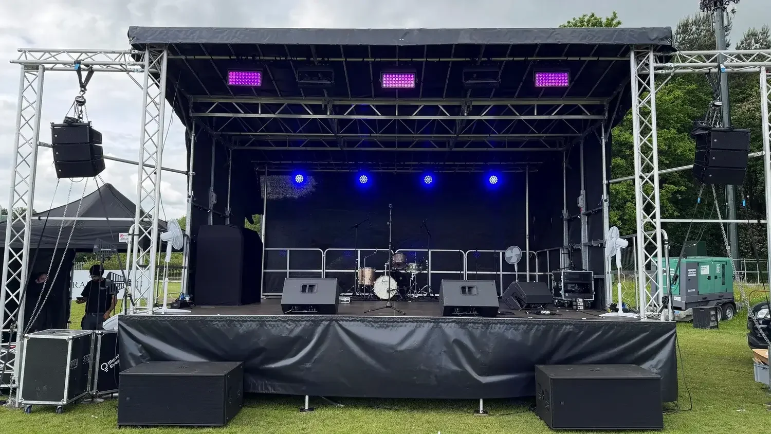 Empty outdoor stage with musical instruments including a drum set and microphone stands, surrounded by sound equipment and purple stage lights, set up for a live performance.