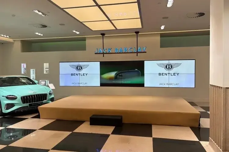 Showroom with a mint green Bentley car and a display wall with digital screens showing the Bentley logo and Jack Barclay branding. Overhead sign reads 'JACK BARCLAY' in blue letters.