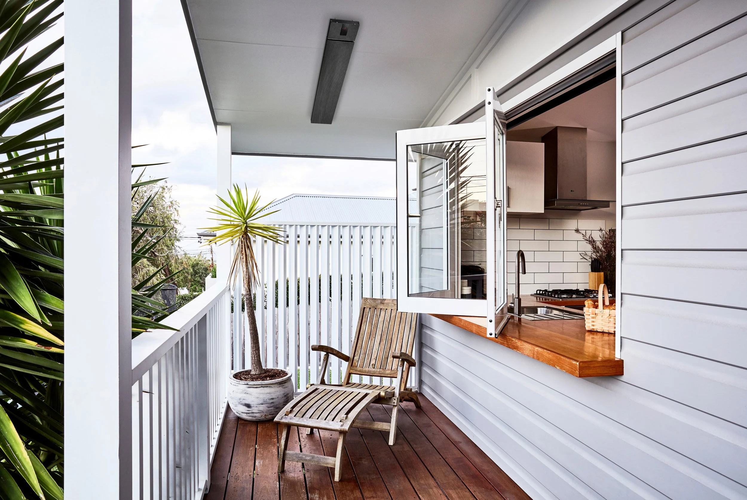 A small outdoor balcony area with wooden flooring, white railings, and a white wooden wall. There are two wooden chairs and a potted palm tree. The balcony is connected to a kitchen interior with white cabinets, a stainless steel sink, and a gas stove. An open window provides a view of the outdoors and a neighboring house's metal roof.