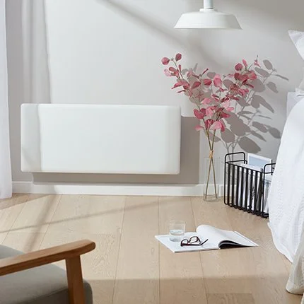 A white radiator, a pink leafy plant, a black wire basket, an open book, and some glasses on a light wood floor in a minimalist room.