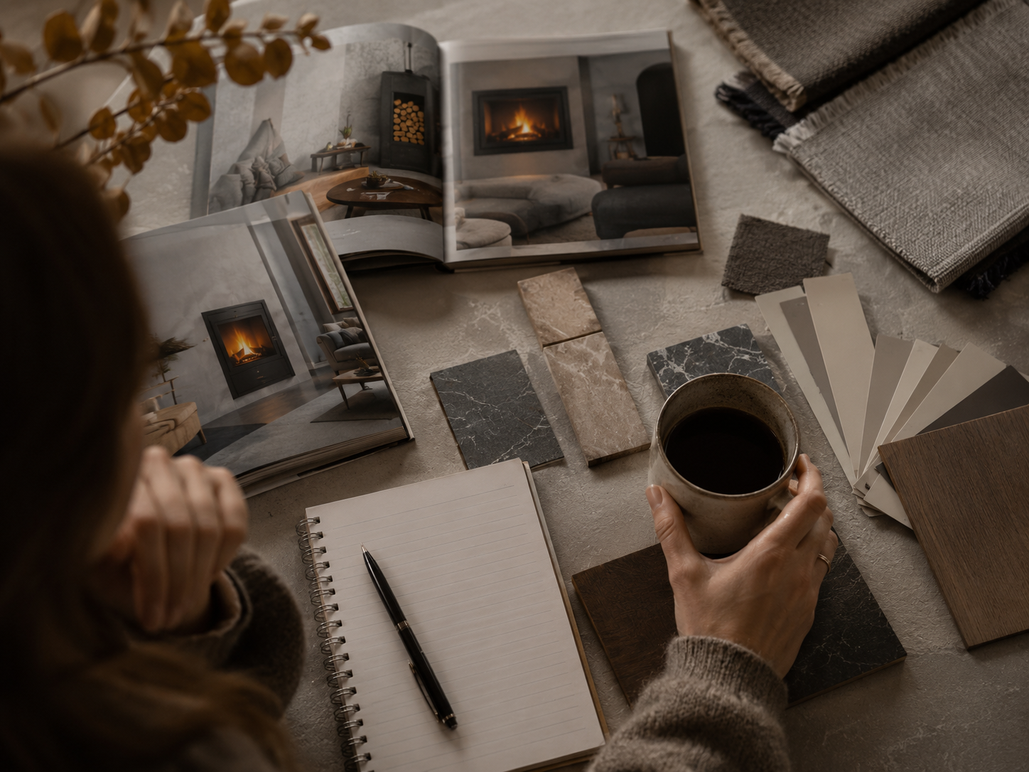 A person holding a mug of coffee surrounded by interior design materials, including fabric swatches, color samples, photographs of living rooms with fireplaces, a notebook, and a pen on a table.