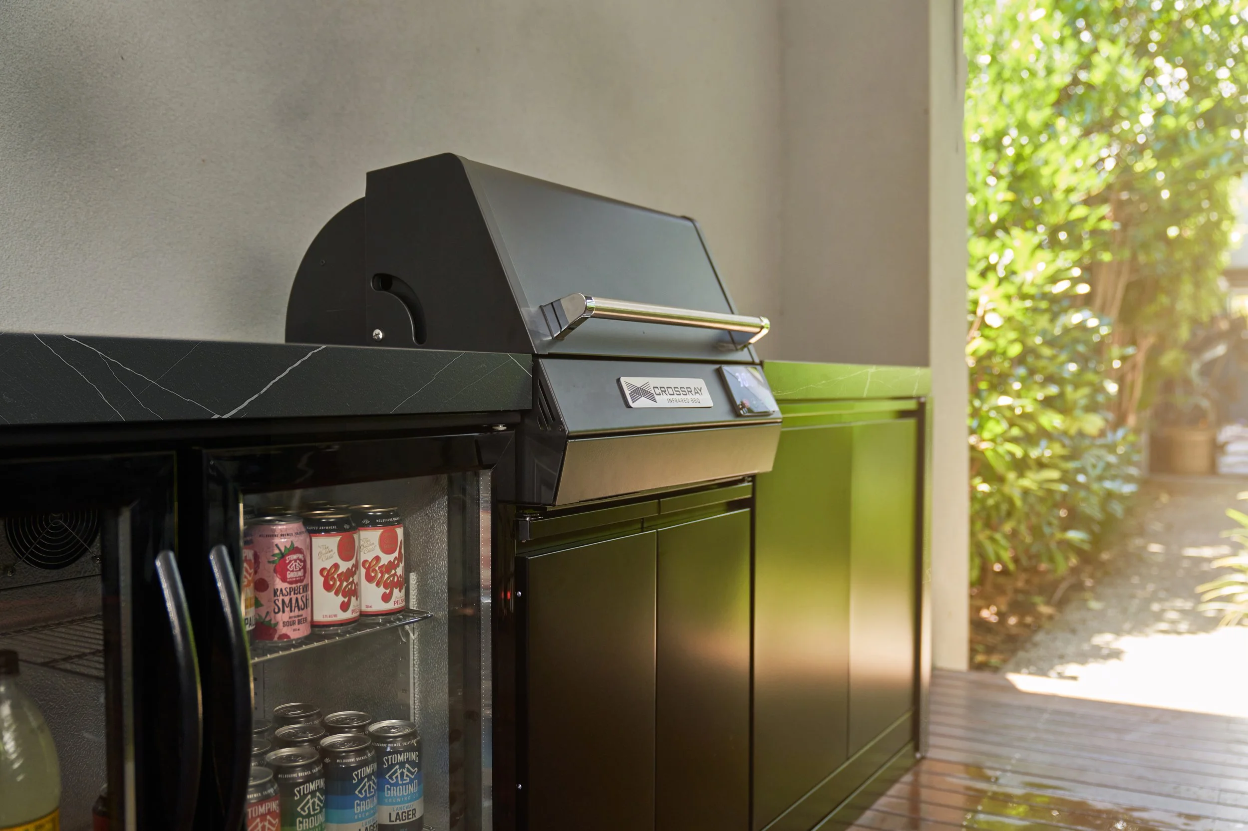 An outdoor kitchen area with a black grill and a clear refrigerator that has cans of soda inside. There are trees and greenery outside with sunlight filtering through.