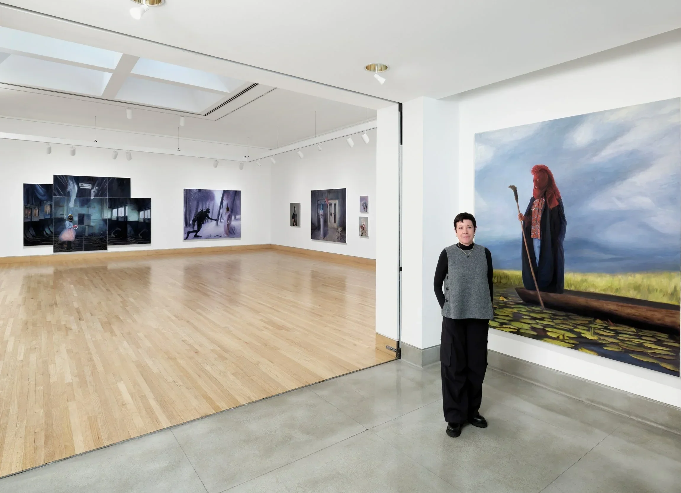 An artist with short dark hair standing in her first solo museum show, dressed in a gray and black outfit, next to a large painting of a figure in a boat with a long staff, set against a cloudy sky backdrop.