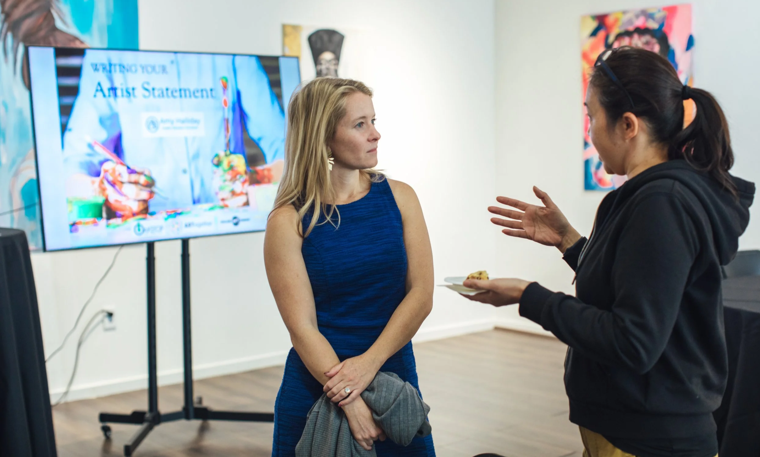 Two women are having a conversation at an art gallery. One woman wears a blue dress and has blonde hair, while the other wears a black hoodie and has dark hair tied back. A large screen in the background displays text about an artist statement. The setting features white walls with colorful artwork hanging.