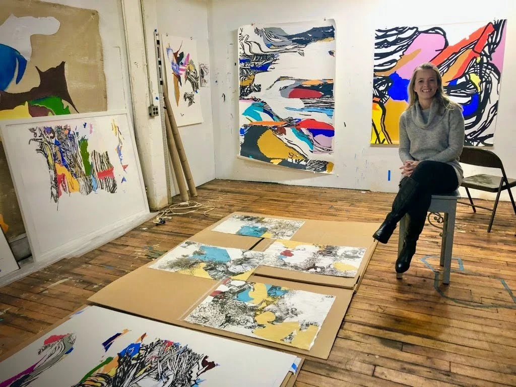 An artist sitting on a stool in her studio surrounded by abstract paintings on the walls and on the floor, which is made of wooden planks.
