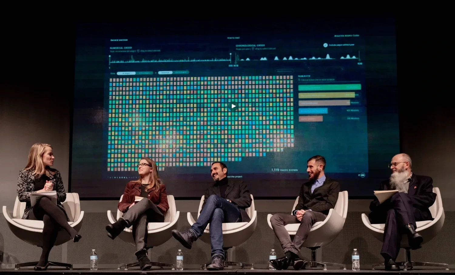 Five people sitting on stage in front of a large screen displaying data visualizations, graphs, and colorful pixelated patterns, engaged in a panel discussion.