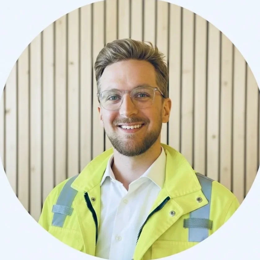A smiling man with glasses and a beard in a white shirt and yellow high-visibility jacket in front of a wooden wall.