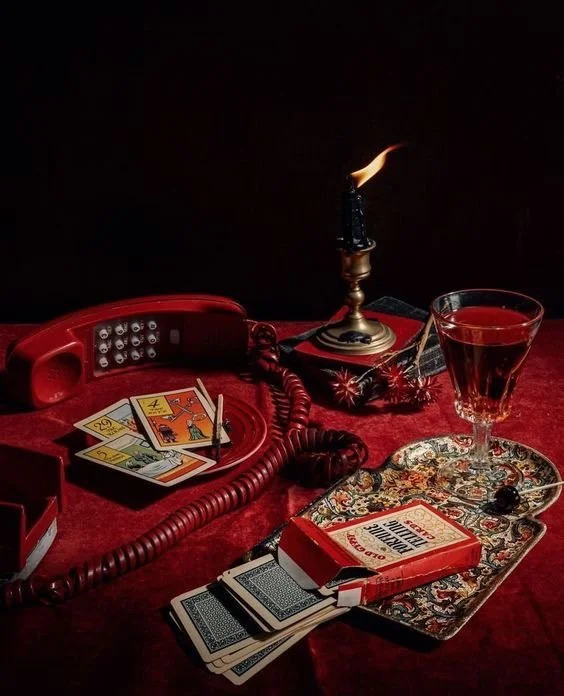 A vintage red rotary phone, tarot cards, a lit candle in a holder, a glass of red wine, a box of matches, and a decorative tray on a dark red surface with a black background.