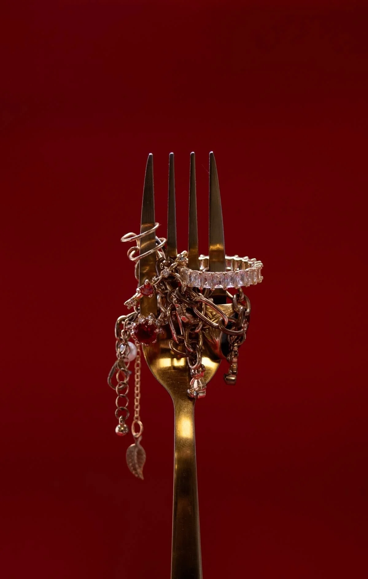 A gold fork with various jewelry items hanging from its tines, against a red background.