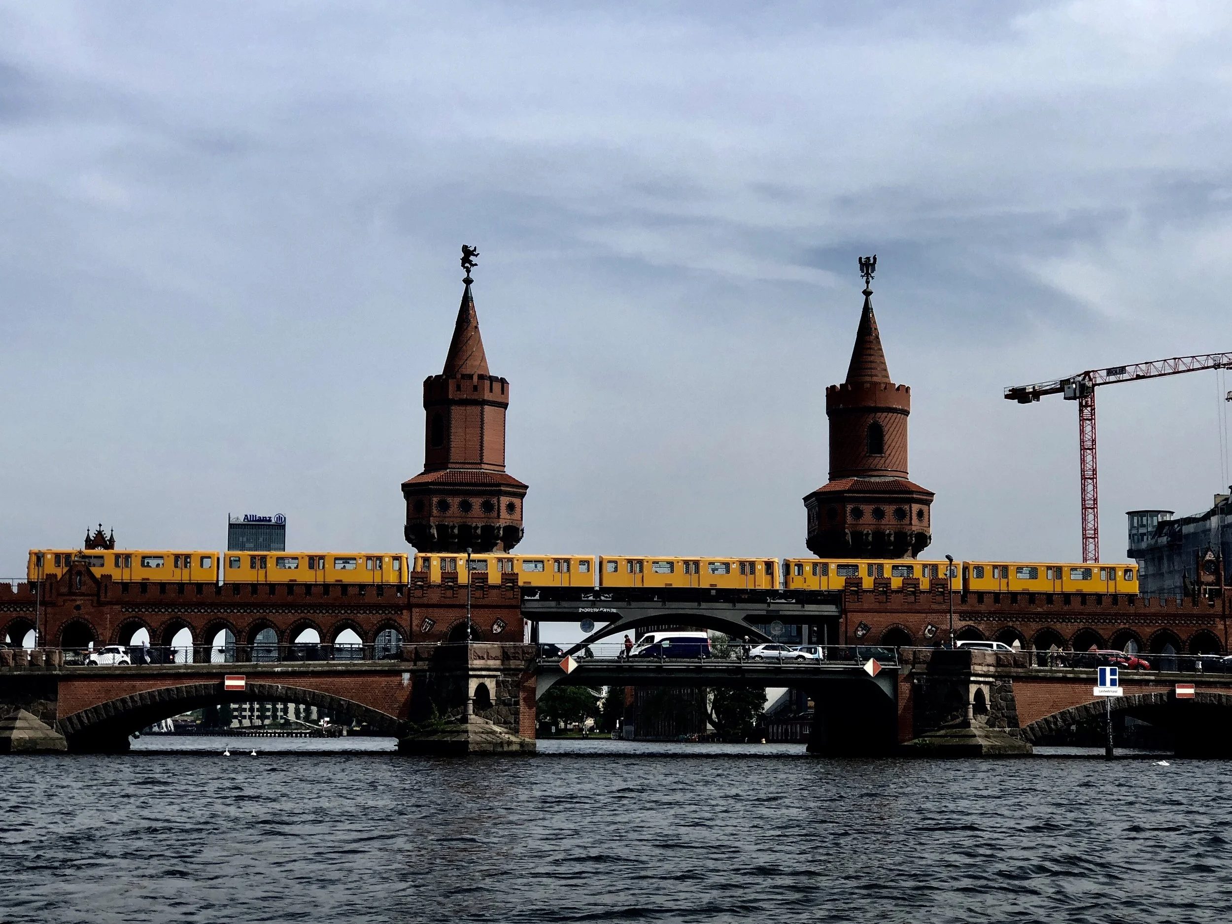 View of a bridge over a river with two prominent red brick towers topped by weather vanes, and a yellow train crossing the bridge. Construction crane and modern buildings are visible in the background, under a cloudy sky.