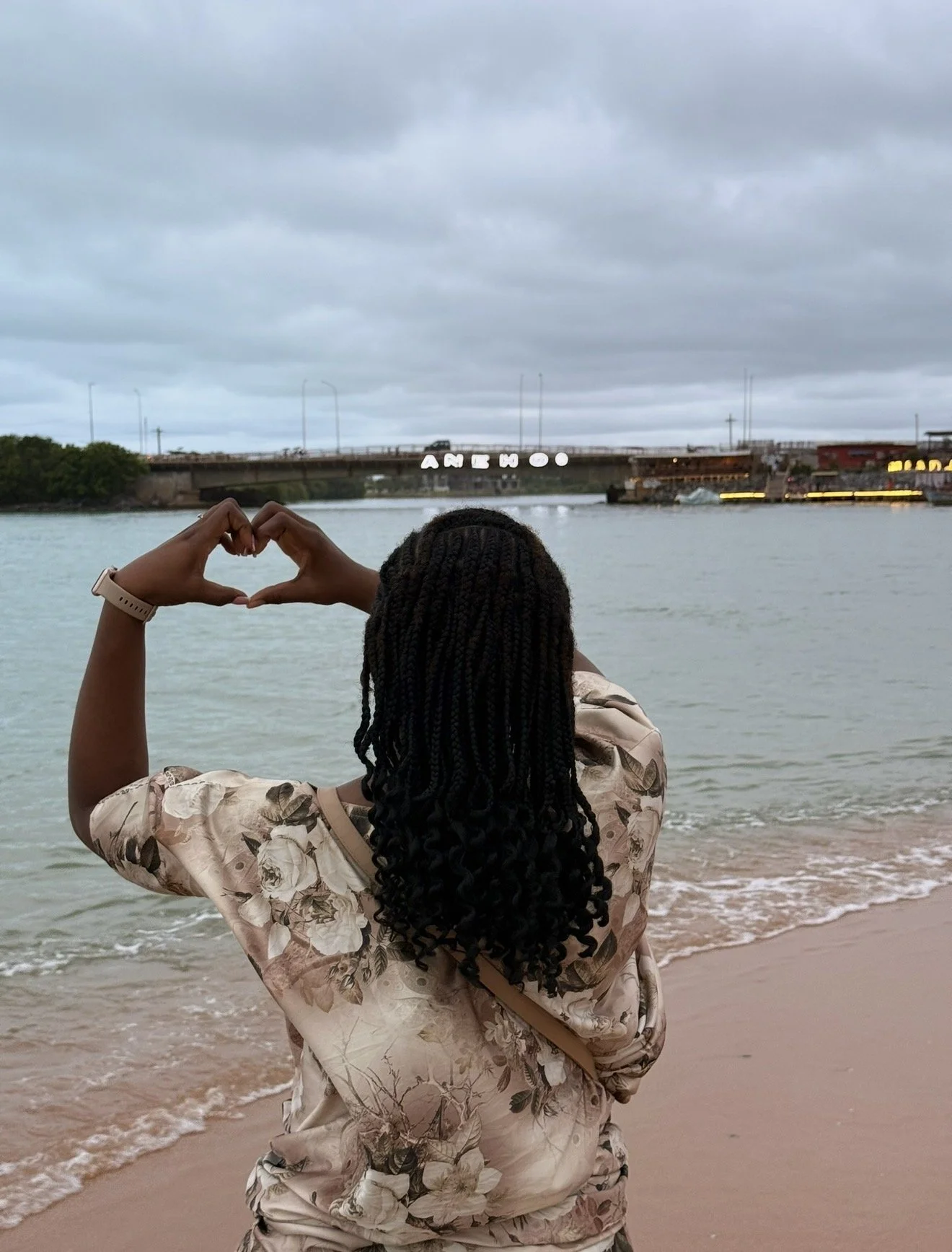 A woman with braided hair wearing a floral shirt faces the water at the beach, making a heart shape with her hands. In the background, a bridge with the word 'Ameoo' lit up spans across the river, under cloudy skies.