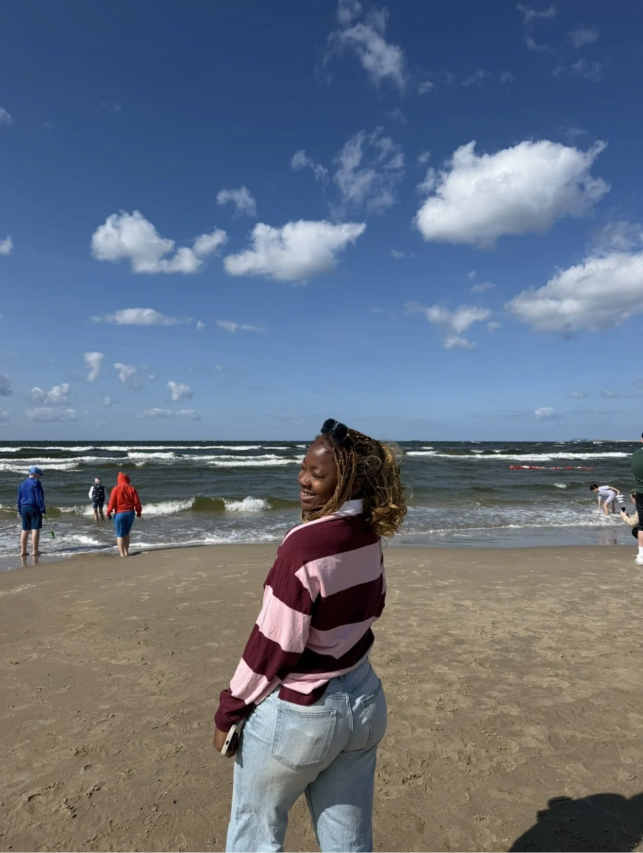 A woman smiling at the beach with the ocean and blue sky with clouds in the background, people wading in the water and playing on the sand.