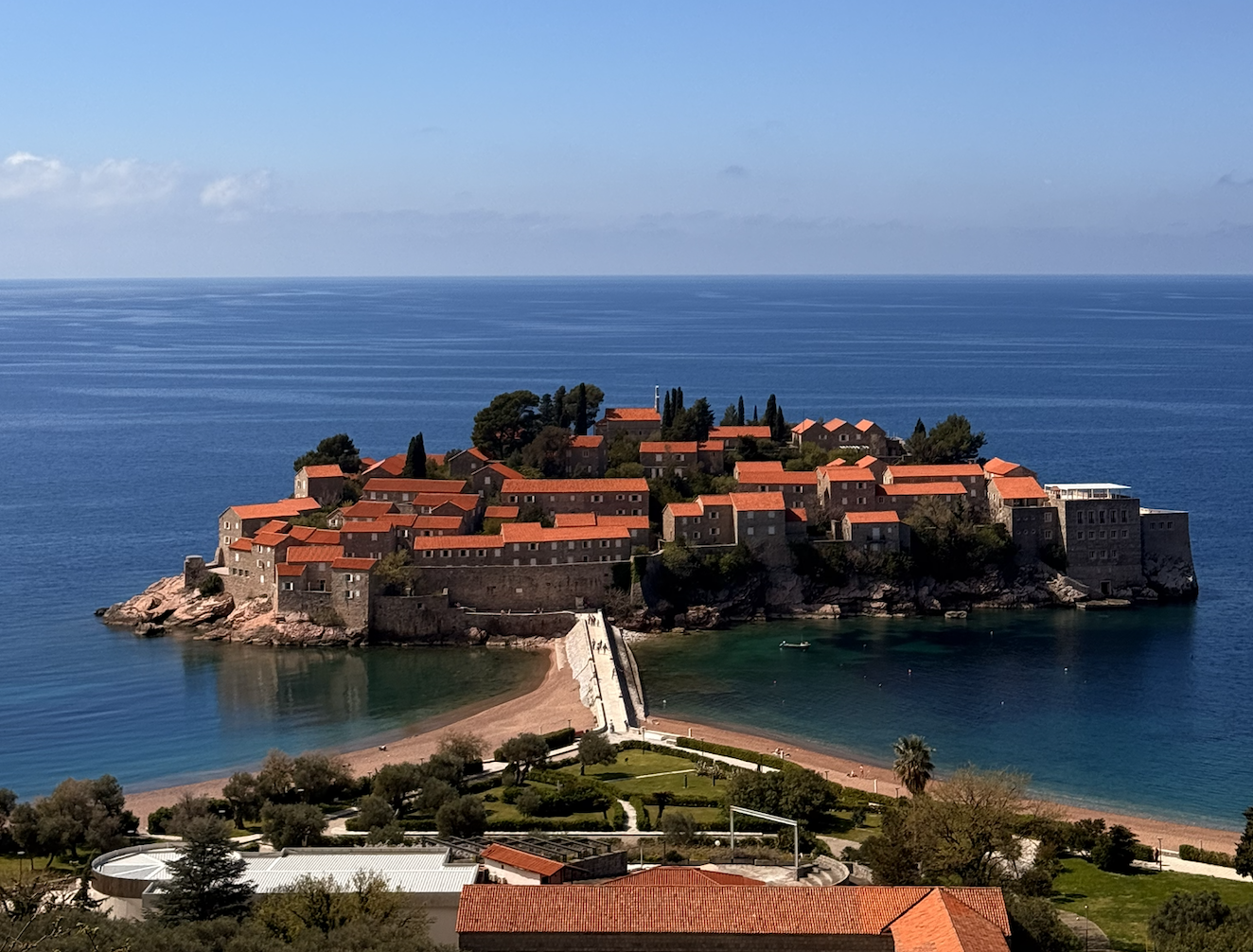 A small island with historic stone buildings and terracotta roofs surrounded by blue sea, with a pier leading to the island, greenery, and a beach in the foreground.