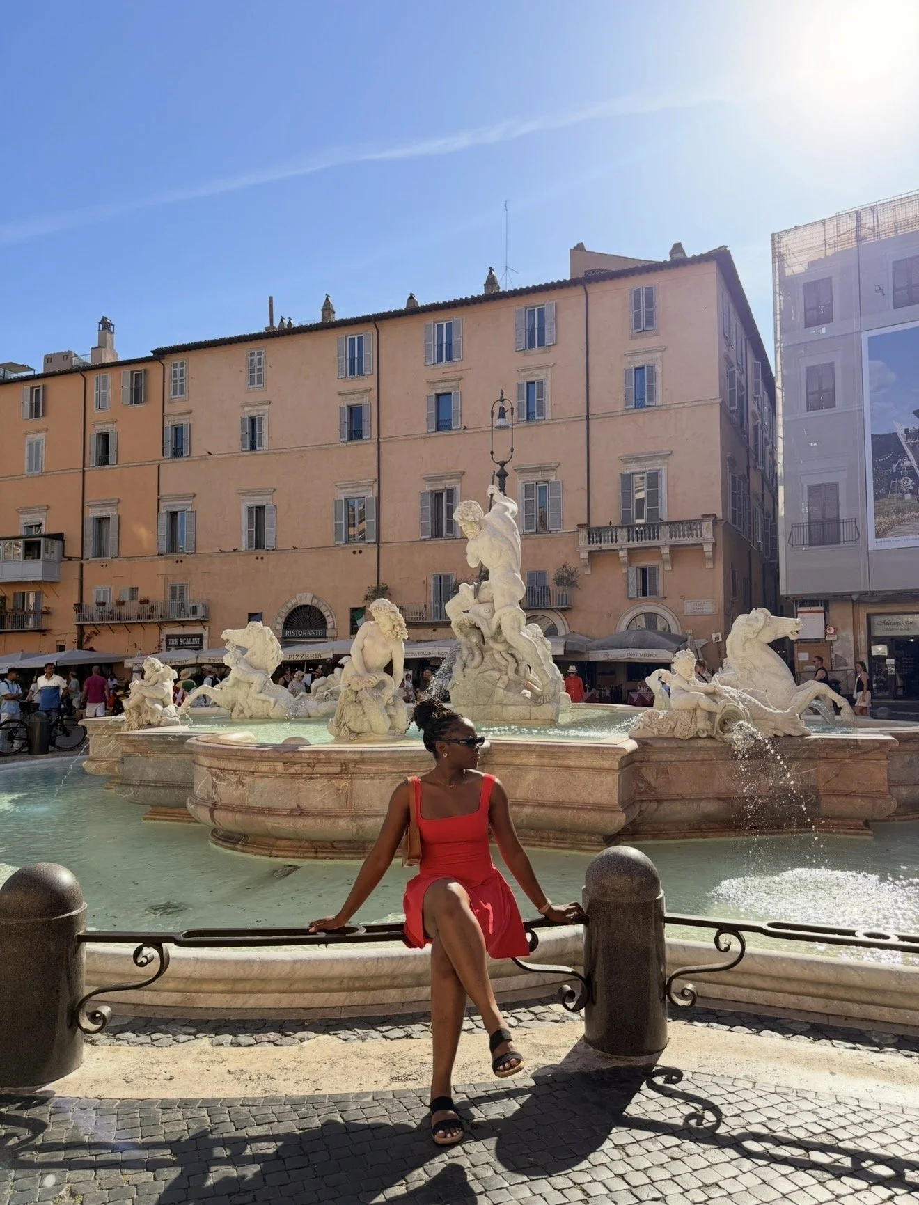 A woman sitting on a fountain with statues of mythological figures in a European city square, peopling and cafes in the background, under a clear blue sky.