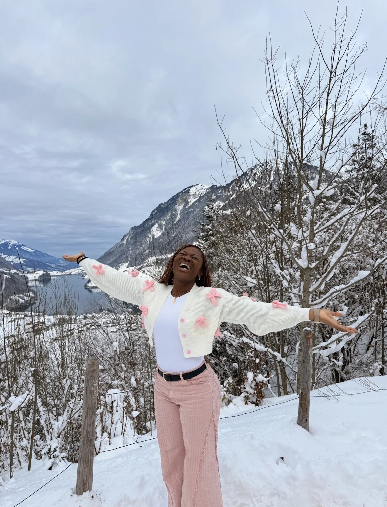 A woman standing outdoors in a snowy landscape, with her arms outstretched and smiling. She is wearing a white cardigan with pink bows, pink pants, and a white top. Snow-covered mountains, leafless trees, and a cloudy sky are in the background.