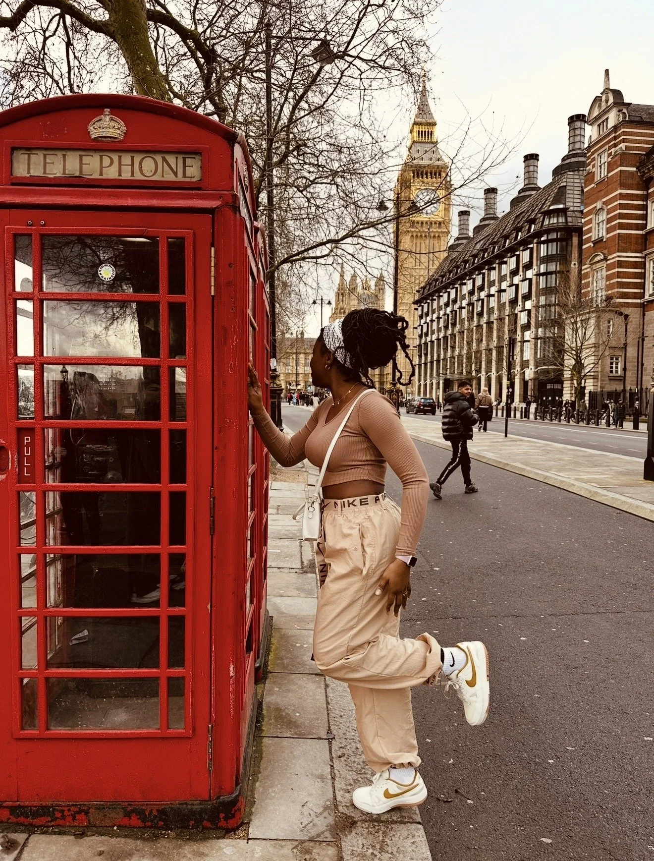 A person with braided hair in a bun, wearing a beige crop top, beige cargo pants, white sneakers, and a white headband, is kneeling on one leg and leaning against a red British telephone booth on a city sidewalk. The background features historic buildings, including the Big Ben clock tower, leafless trees, and a few pedestrians walking along the street.