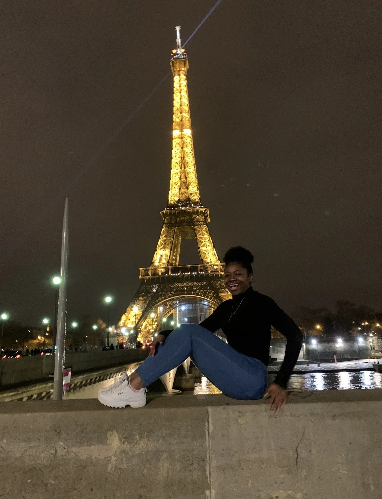 A woman in black top and blue jeans sitting on a ledge in front of the Eiffel Tower lit up at night in Paris, France.