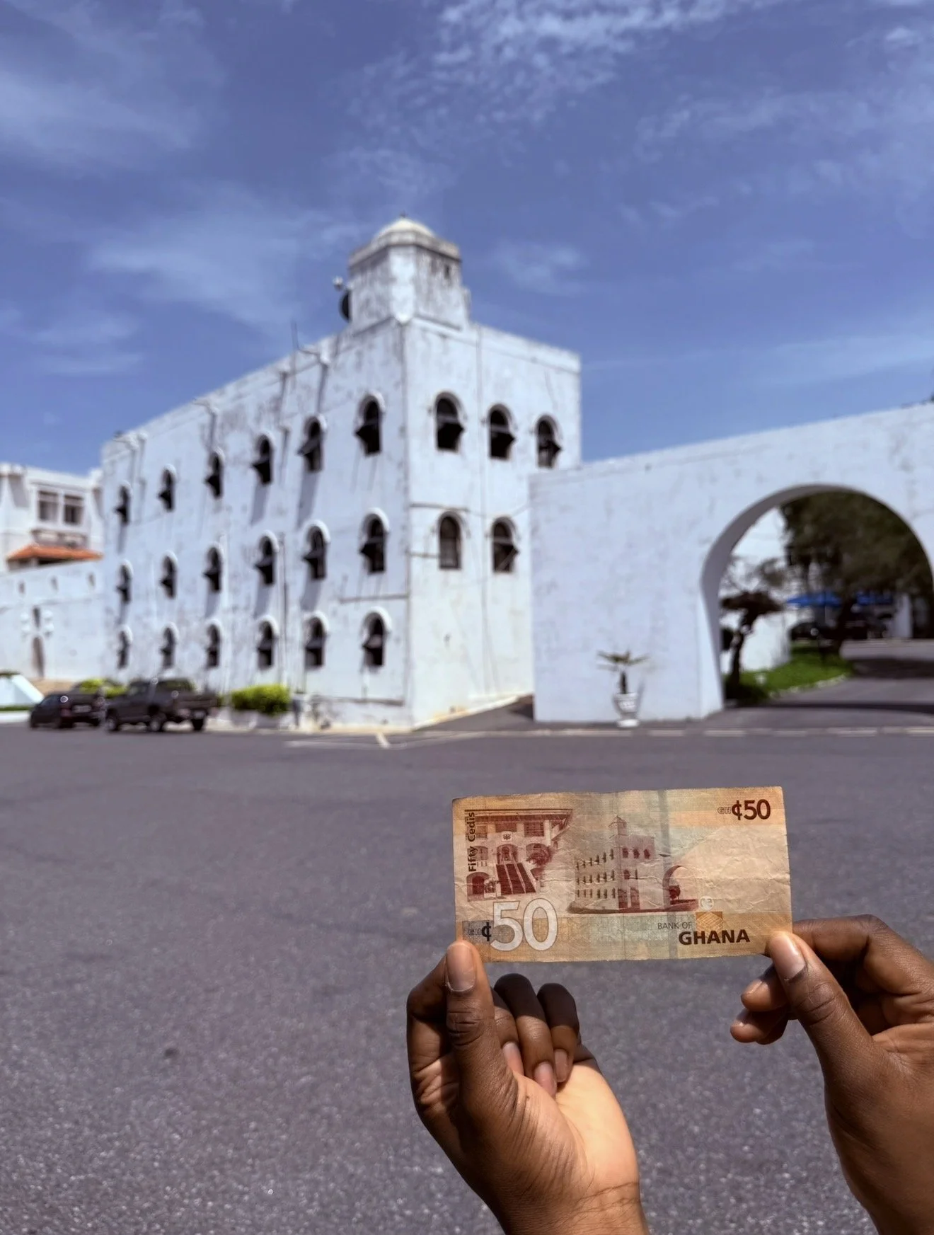 A person holding a 50 Ghanaian cedis banknote in front of a white historic building with a clock tower and an arched entrance in Ghana. The building is under a clear blue sky.