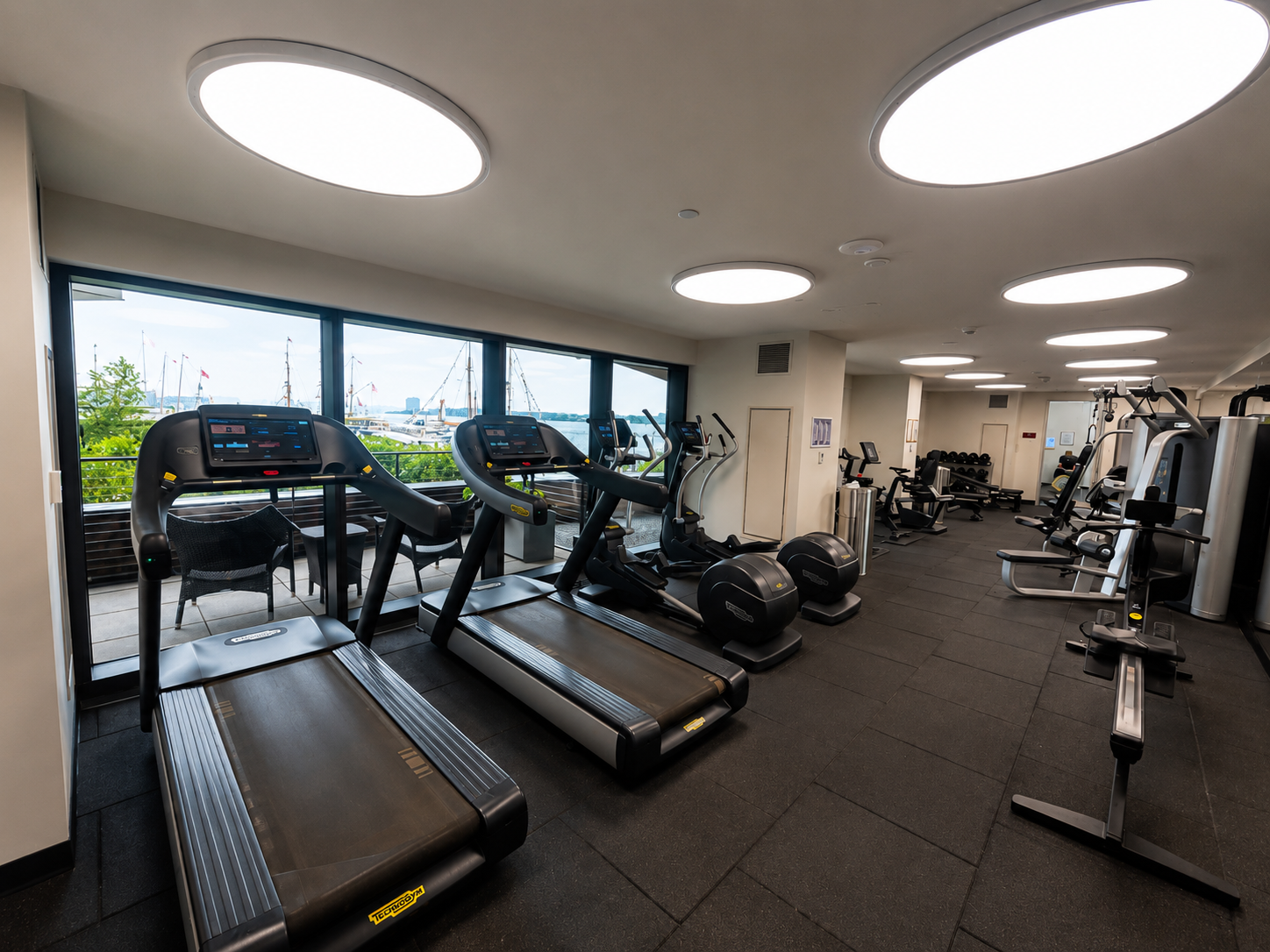 Modern gym with treadmills and exercise equipment near large windows overlooking waterfront with boats.