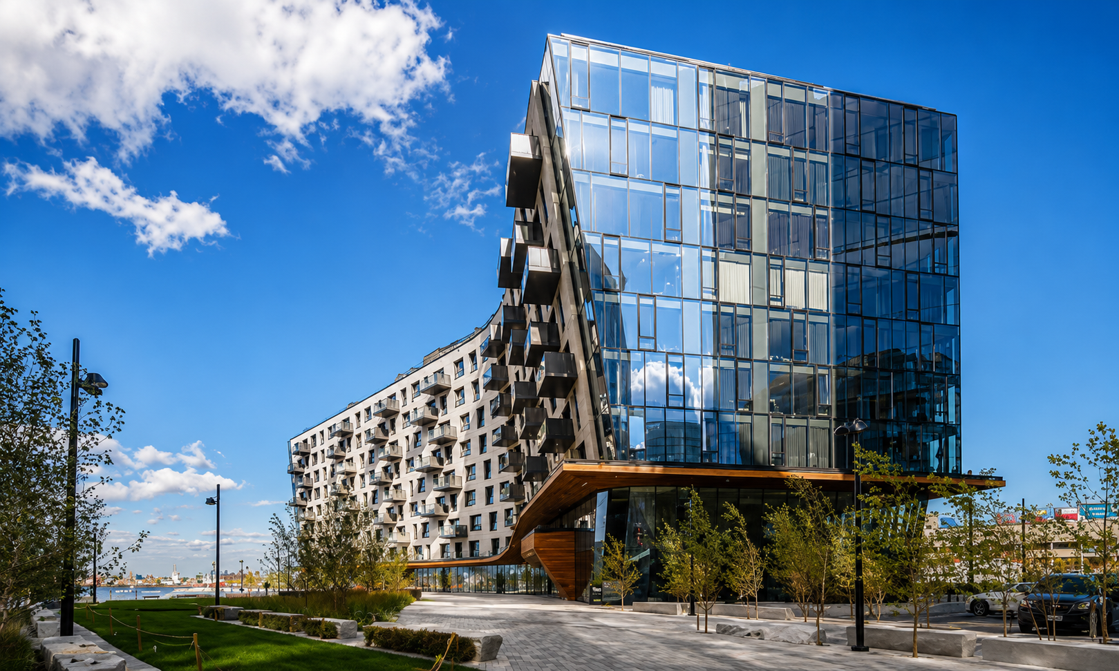 Modern mixed-use building with glass and concrete facade, reflecting blue sky and clouds, and surrounded by a landscaped area with trees and street lamps.