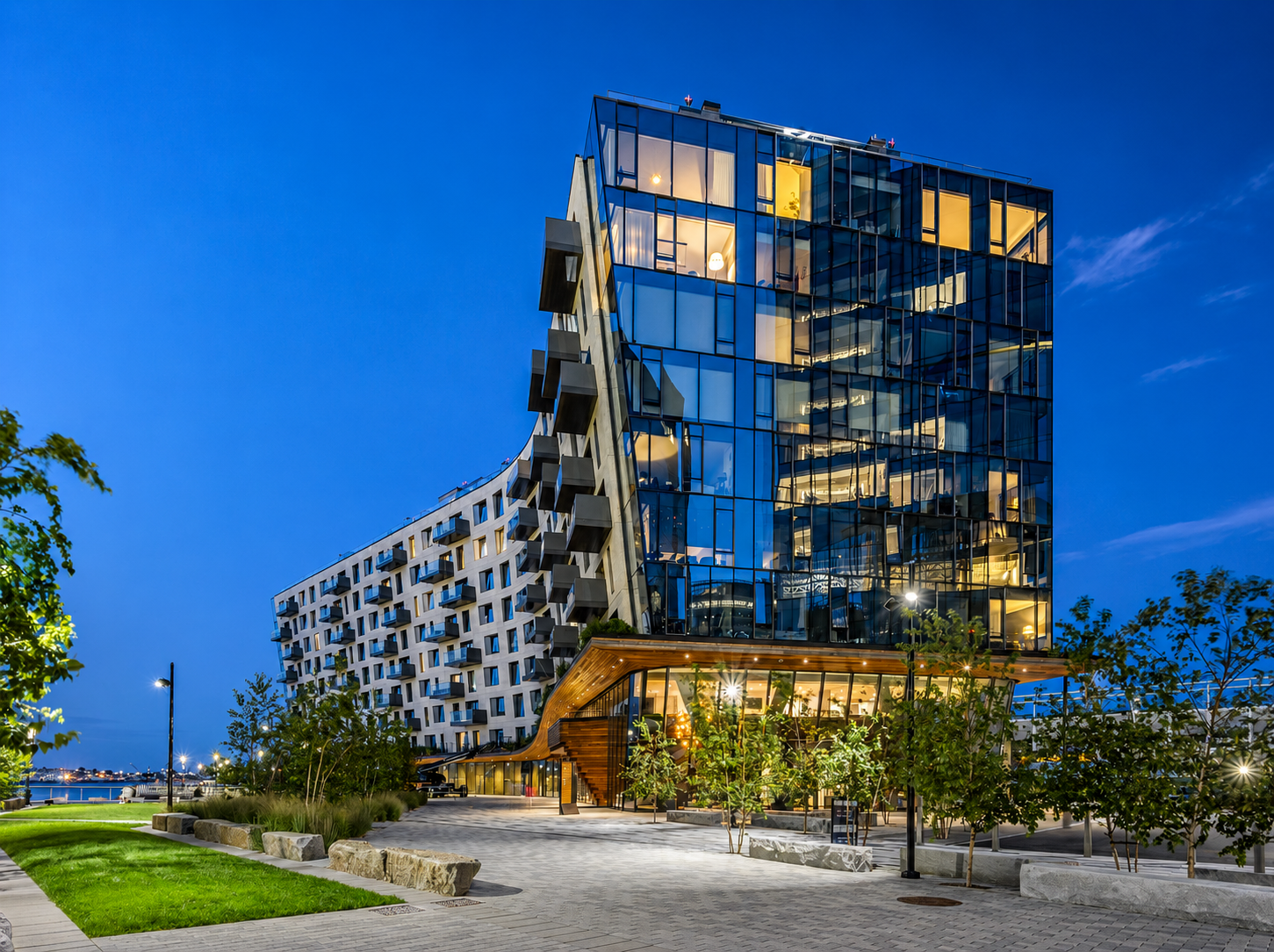 Modern multi-story building with extensive glass windows and unique architectural design, illuminated at dusk, with trees and a paved walkway in the foreground.