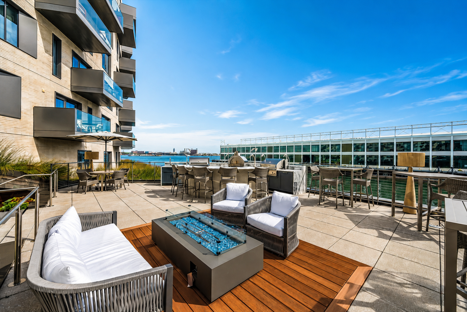 Rooftop patio with outdoor seating, including a sofa and armchairs around a fire pit, bar stools at a bar area, and a grill, with a view of the water and a building with glass windows under a clear blue sky.