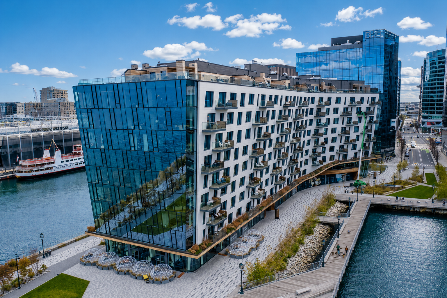 Modern waterfront building with reflective glass facade and small balconies, situated along a river with a park and dock, in a city with other high-rise buildings under a blue sky with scattered clouds.