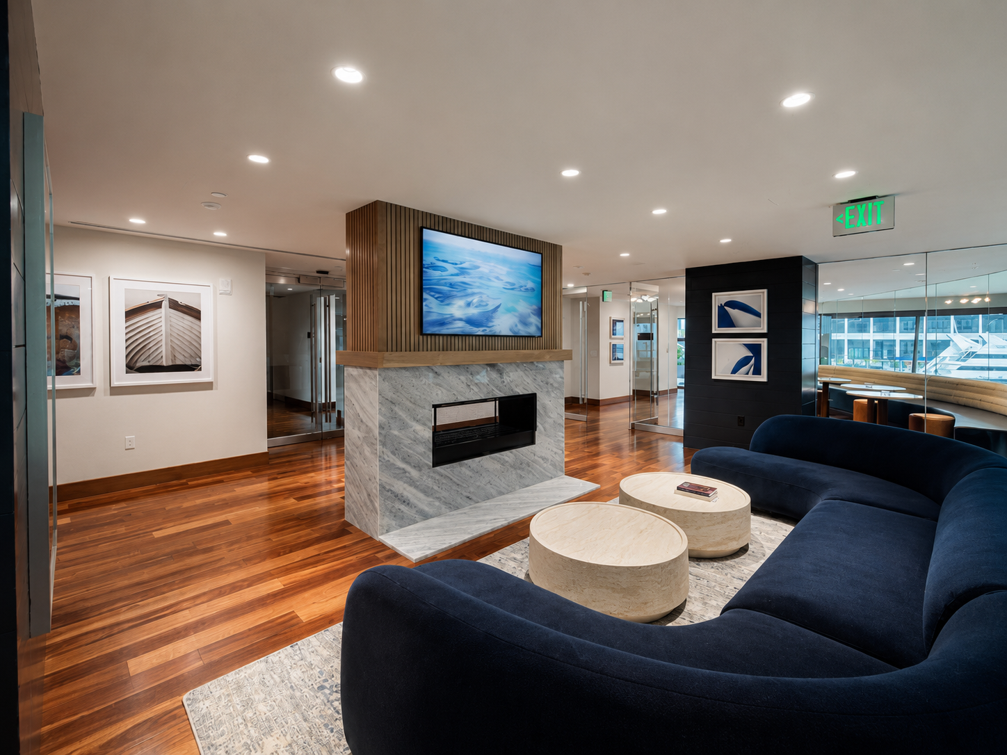 Modern hotel lobby with a dark blue curved sofa, round marble coffee tables, a fireplace with a marble surround, and a wall-mounted TV displaying abstract ocean art. The space features wooden flooring, framed artwork, and an emergency exit sign.