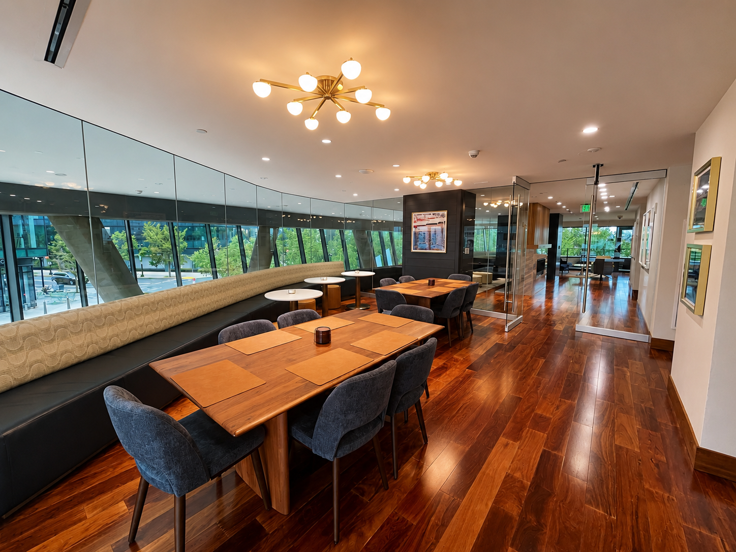 Modern restaurant or cafe with wooden floors, large windows, and a mix of wooden and upholstered seating. Decor includes hanging lights and framed artwork on the walls.