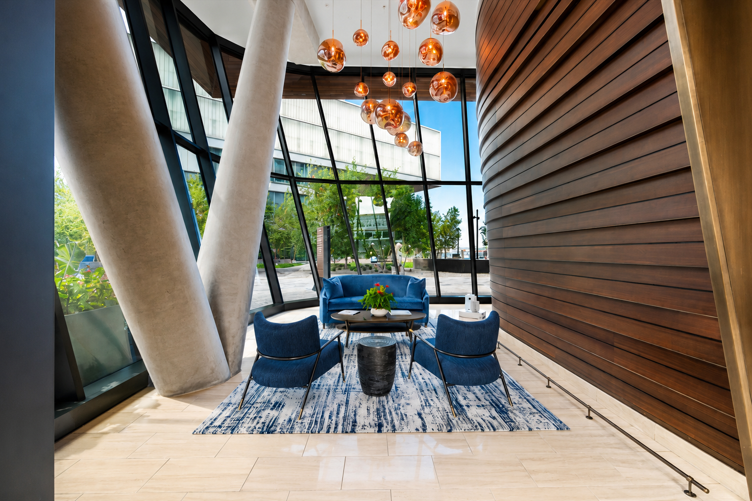 Modern hotel lobby with seating area, including a blue sofa, two blue chairs, a patterned rug, a small black table with a plant, and a large window with a view of trees and buildings outside. A chandelier with copper-colored spherical lights hangs from the ceiling, and the walls feature wood paneling.
