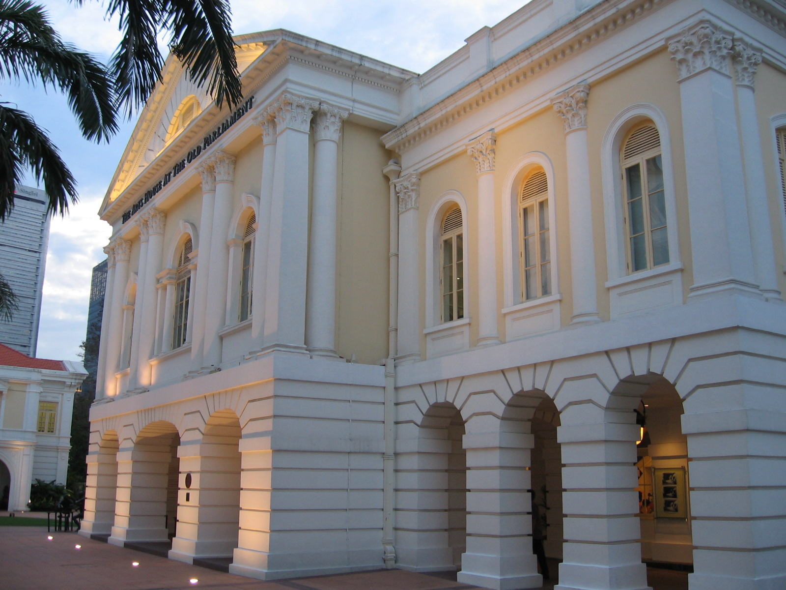 A white, neoclassical-style building with tall columns, arched windows, and decorative moldings, lit softly at dusk with palm trees nearby.