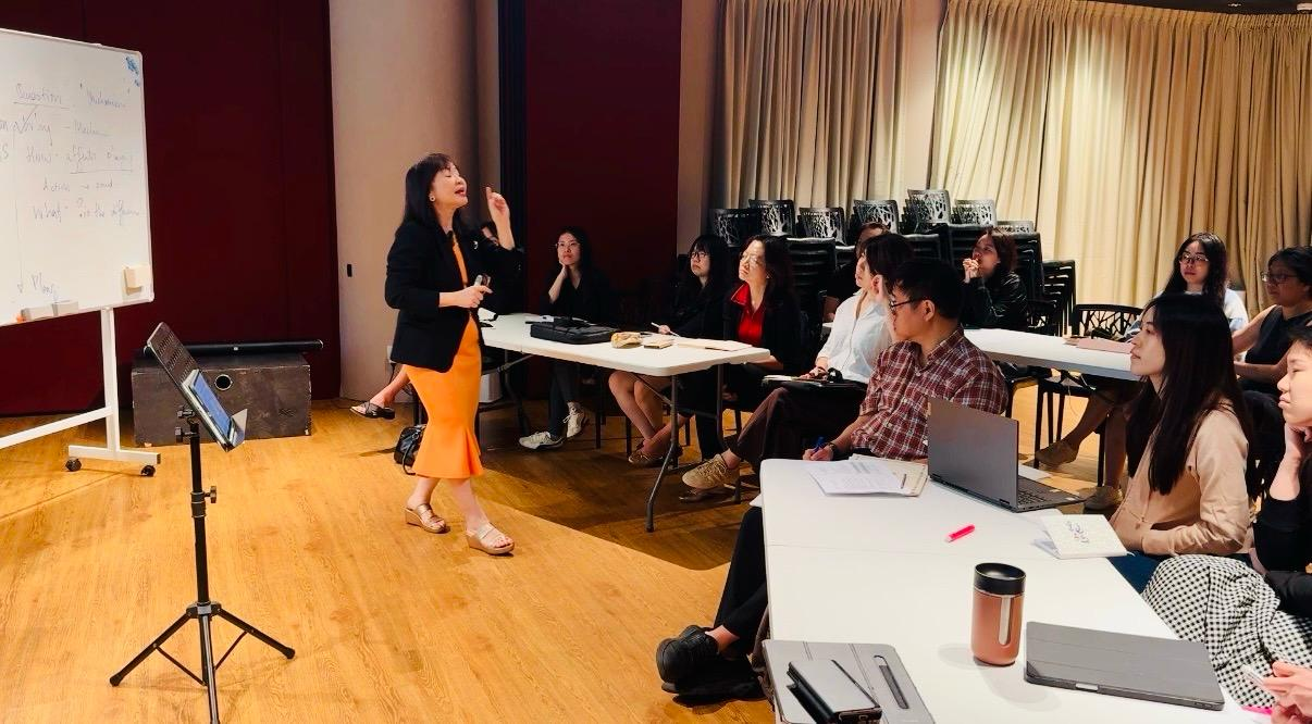 A woman in an orange dress and black blazer presenting to a group of seated young adults in a classroom or conference room setting.