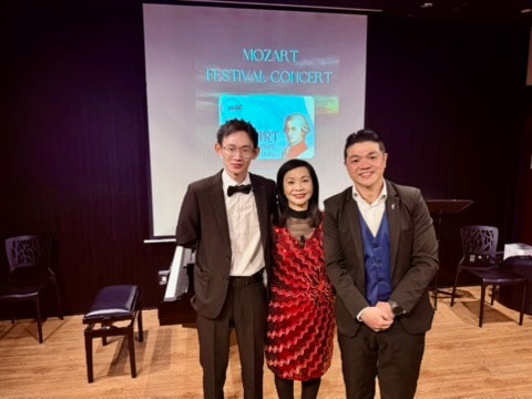 Three people standing in front of a presentation screen at a concert event, with two men and one woman dressed in formal attire.