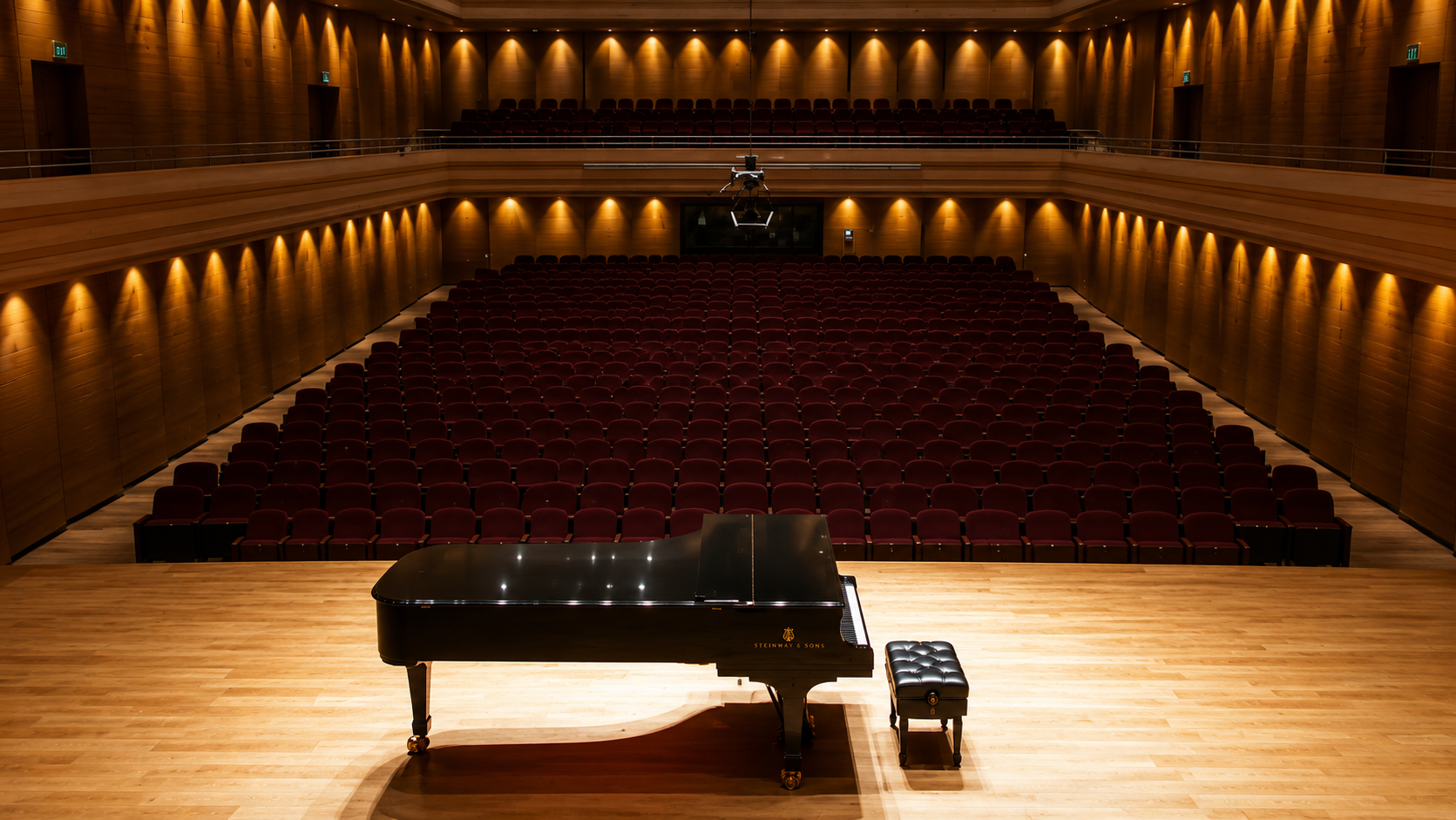 Empty concert hall with a black grand piano on stage and rows of red seats, wooden walls, and warm lighting.