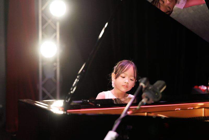 Young girl playing the piano on stage with bright lights behind her.