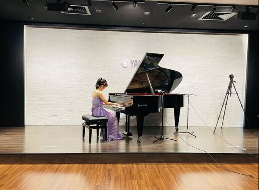 A woman in a purple dress playing a grand piano on a stage with a white background and wooden floor, with a camera on a tripod nearby.