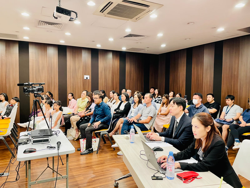 Audience members attending a presentation or conference in a room with wood-paneled walls, several people seated at tables with laptops, water bottles, and notebooks, some wearing face masks.