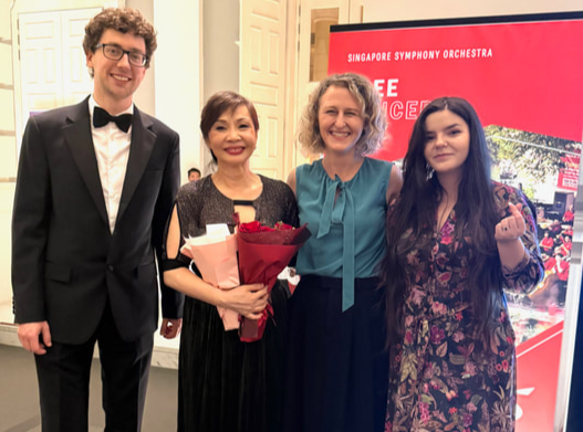 Four people standing together indoors, dressed formally. The woman second from the left is holding a bouquet of red flowers. Behind them is a pink and red sign that says 'Singapore Symphony Orchestra'.
