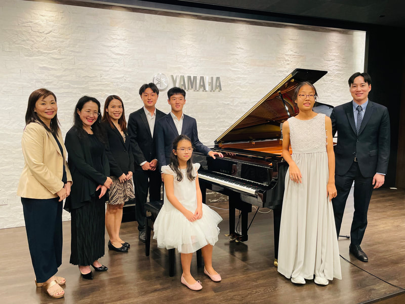 Group of seven adults and two children posing in front of a Yamaha grand piano, indoors, with a white brick wall background.