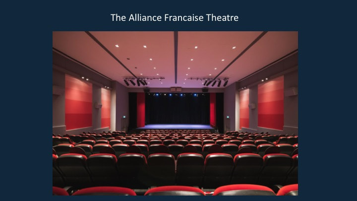 Interior of the Alliance Francaise Theatre with rows of red and black seats facing a stage with a black curtain, pink and red wall panels, and ceiling lighting.