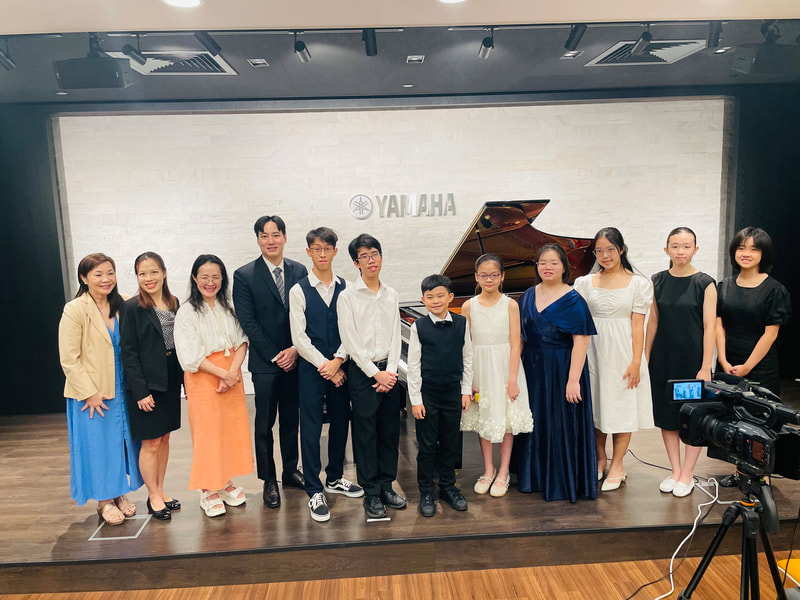 A group of 13 people standing on a stage, with a Yamaha logo and a grand piano behind them in a music performance or recital setting.