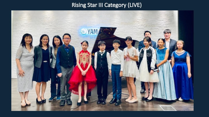 Group of young girls and boys posing on stage after a performance, with a woman in a red dress and a man in a suit, at the Rising Star III Category live event.