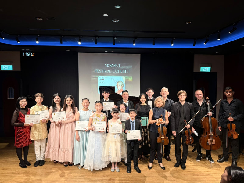 Group of children and adults holding certificates and musical instruments on stage after a concert, with a screen behind displaying 'Mozart Festival Concert'.