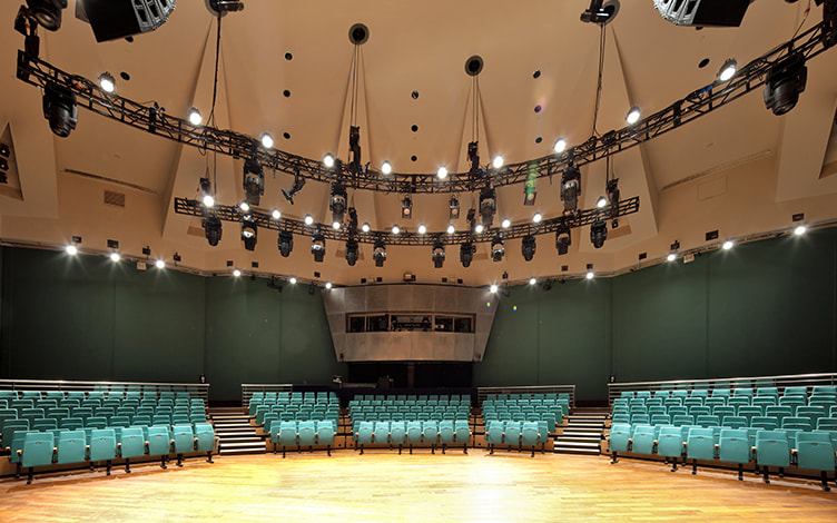 An empty theater or auditorium with teal-colored seats, a wooden stage, and hanging stage lights from the ceiling.