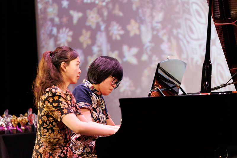 Two women playing the grand piano together on stage during a performance.