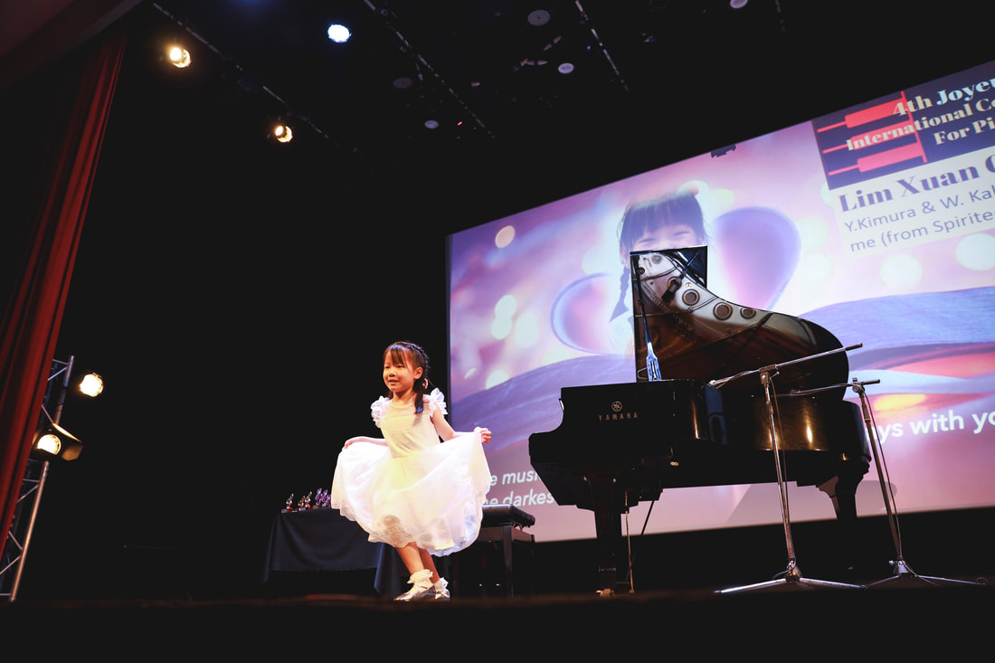 A young girl in a white dress stands on stage near a black grand piano, with a large screen behind her displaying colorful graphics and text.